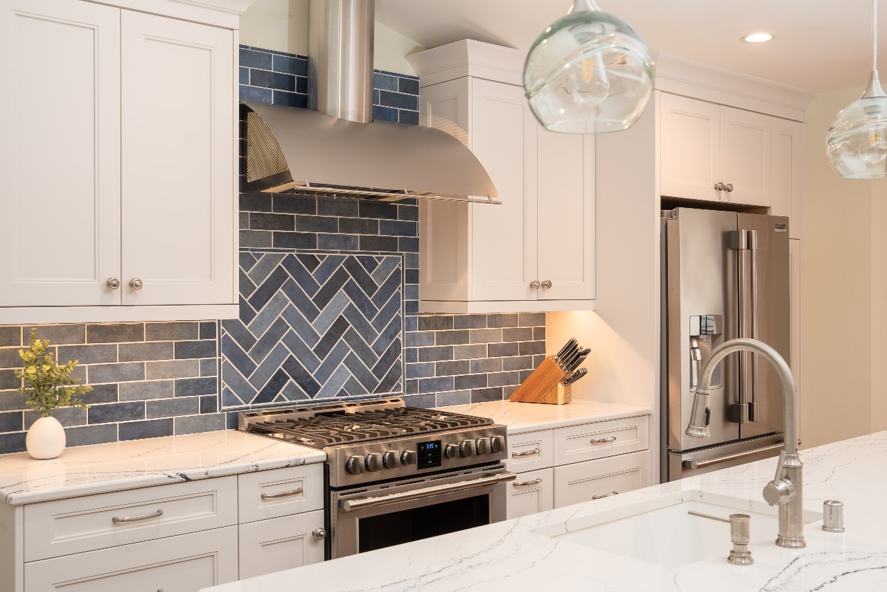 White cabinets, blue subway tile backsplash and stainless steel refrigerator, oven and range hood with the island in the foreground in a remodeled custom kitchen.